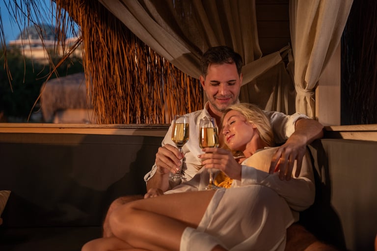 A man and woman share a tender moment with champagne glasses in a cozy, dimly lit cabana overlooking a beach at dusk