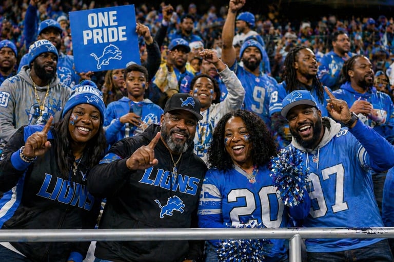 Detroit Lions fans cheering in blue jerseys and hats, holding a One Pride sign at a stadium game
