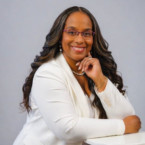 Professional portrait of a smiling woman with long dark wavy hair, wearing glasses, a white blazer, pearl necklace, and silver bracelets, posing against a gray background